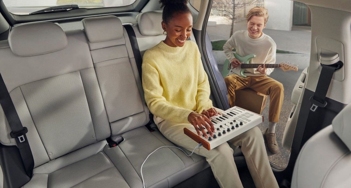 Two women are sitting in the front row of a The all-new KONA with their seats folded back, watching the sunset. The woman in the front passenger seat is holding her smartphone, and the woman in the driver's seat is holding her take-out paper cup in her left hand, with her arms crossed.