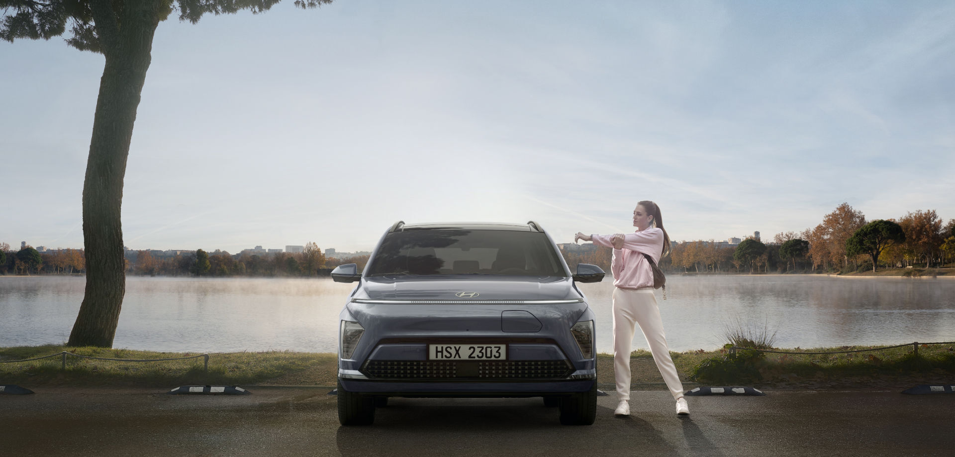 The front view of a The all-new KONA Electric standing in a parking lot by the lake. On the left side, a woman in sportswear is doing warm-up exercises.