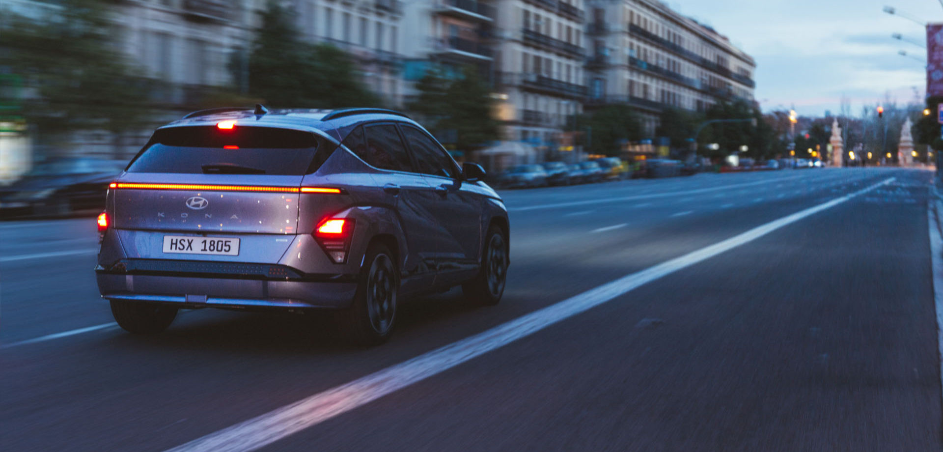 Rear view of The all-new KONA with red taillights running on a darkened city road.