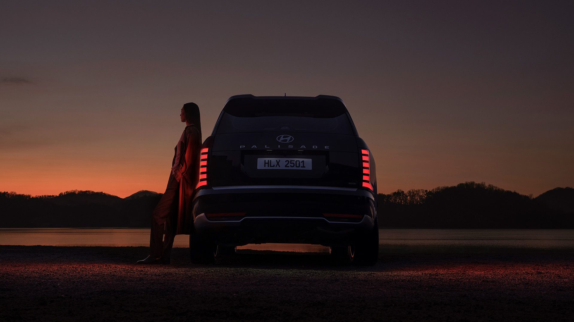 A woman leaning on the side of the all-new PALISADE, gazing at the sunset