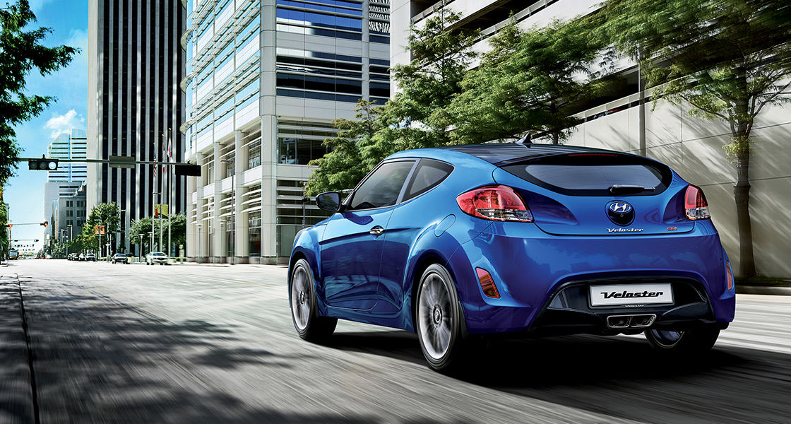Left side rear view of blue Veloster on the road with the buildings beside