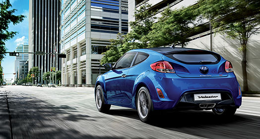 Left side rear view of blue Veloster on the road with the buildings beside