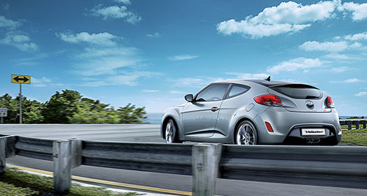 Left side rear view of dark gray Veloster driving on the road on a clear day
