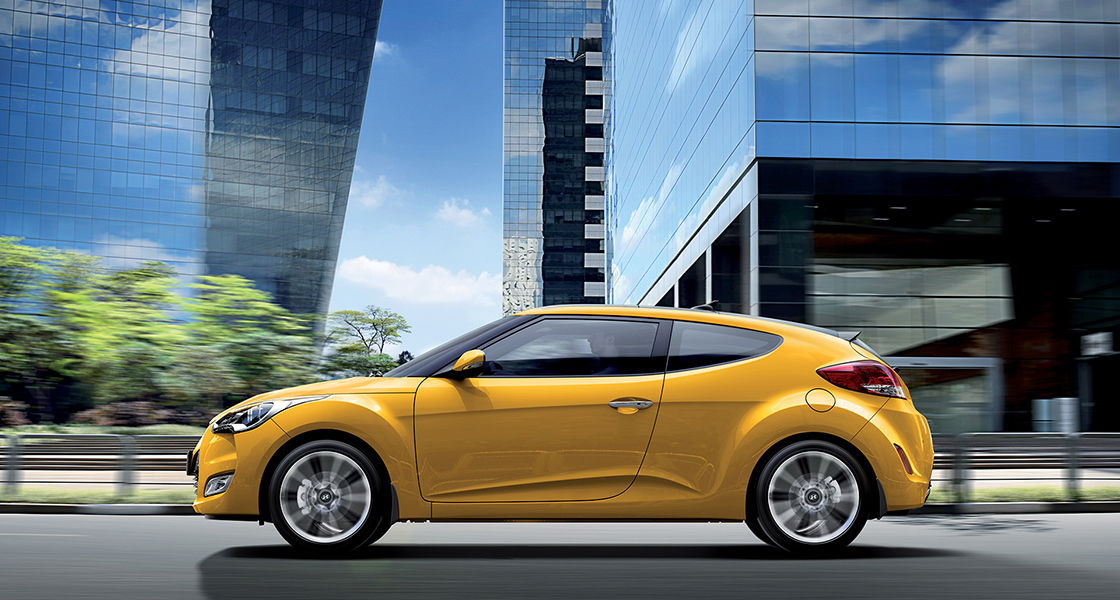 Left side view of yellow Veloster behind buildings