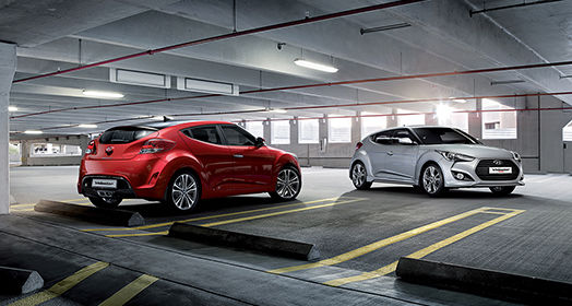 One side rear view of red veloster and side view of gray veloster parked in indoor parking lot