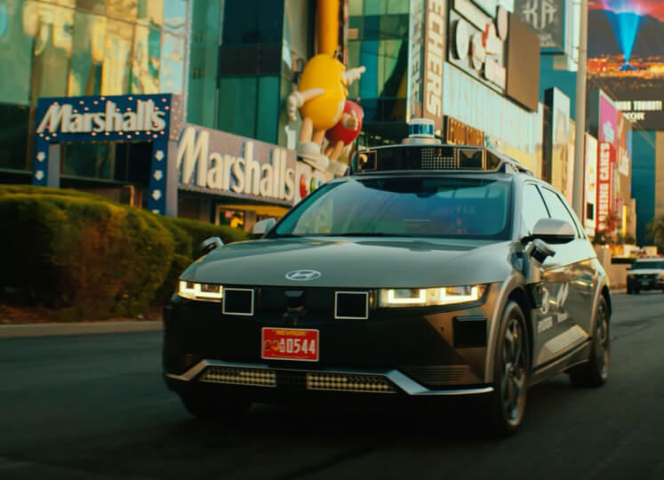 Autonomous driving : A gray IONIQ 5 robotaxi driving in traffic in downtown Las Vegas. The sensor is clearly visible on the roof and the parametric headlights are lit up.