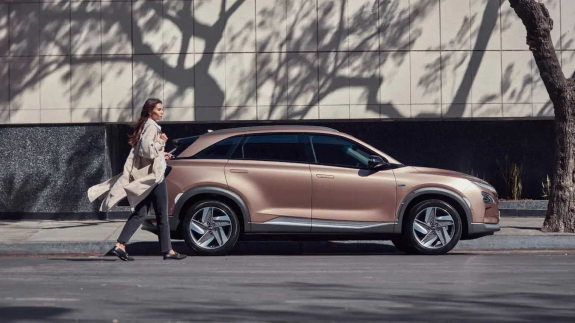 A woman standing next to a Hyundai NEXO hydrogen fuel cell vehicle in an urban environment.