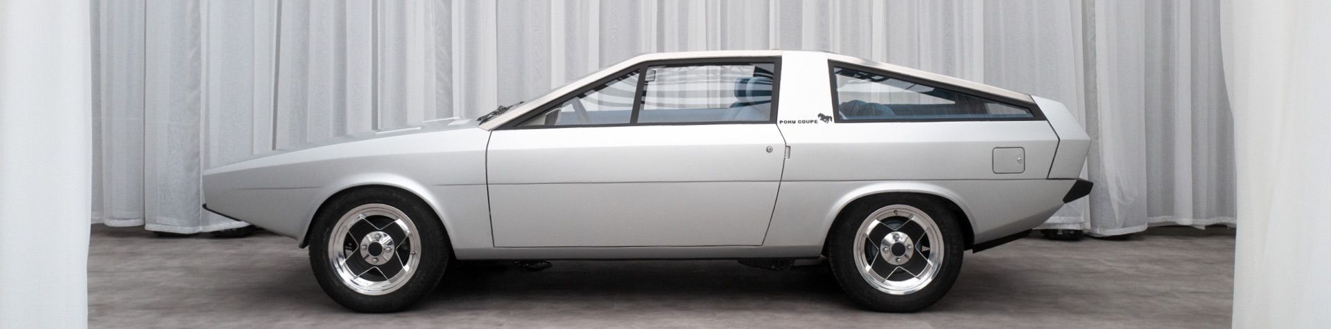 은색 휠 커버와 함께 회색으로 복원된 포니 쿠페 콘셉트 카의 옆모습이 하얀색 커튼으로 장식된 무대에서 선보이고 있다.