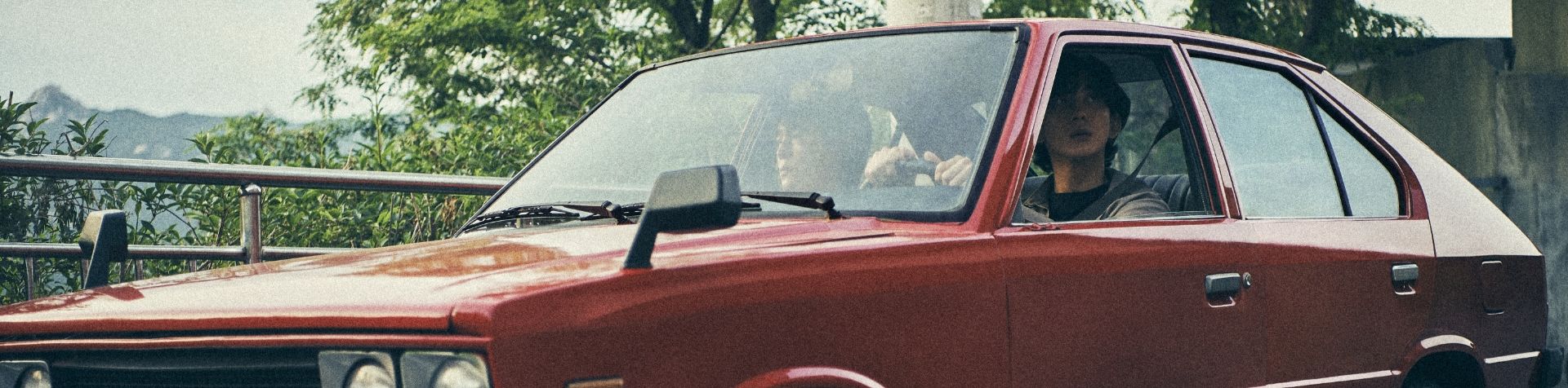 Two Korean men driving in a red 1970s Pony along a tree-lined road with mountains on the horizon.