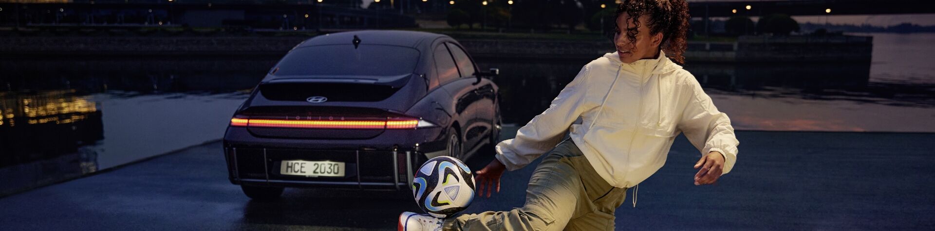 Night-time image of woman in casual street attire from modern times volleying a football in front of a Hyundai IONIQ 6 along a waterfront.