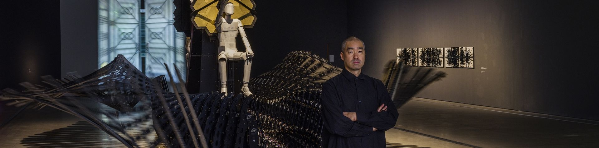 Artist Choe U-Ram standing in front of his Little Ark exhibition. He is wearing a black shirt and has his arms folded across his chest. Behind him the Little Ark piece is moving and there is a white human sculpture sitting above it with a hexagonal gold piece set behind it.