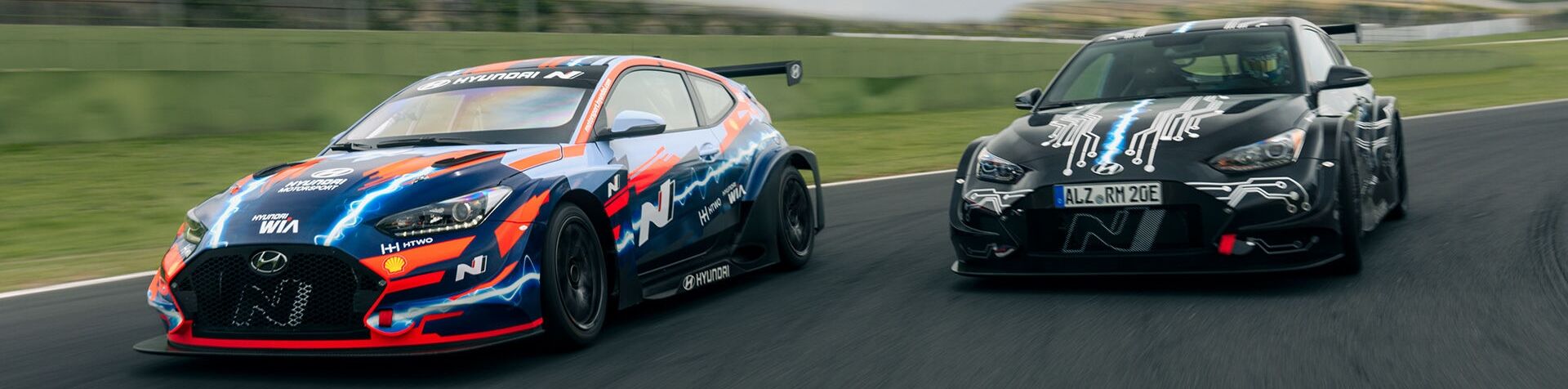 Two Hyundai N RM (Racing Midship) sportscars racing down a racetrack. The car on the left is red and blue and the car on the right is black.