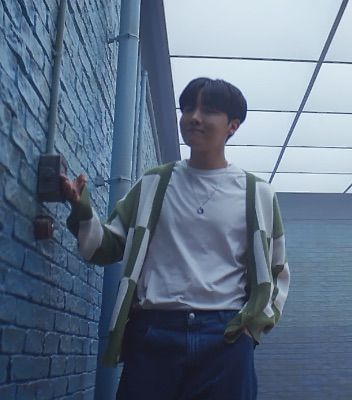 BTS member J-Hope stands in a room with white brick walls and goes to flip the light switch.