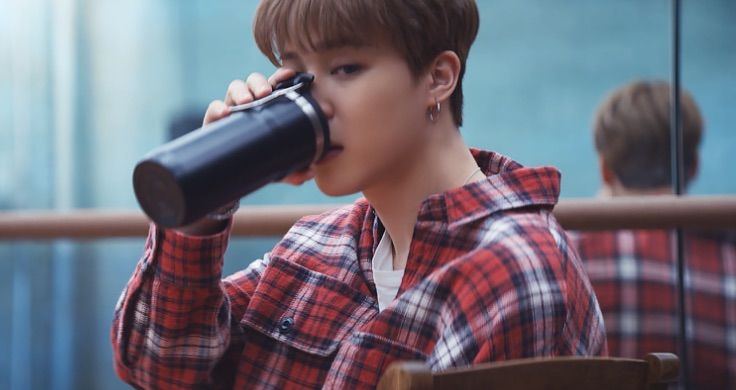 BTS member Jimin wearing a red, blue and white chequered flannel shirt takes a sip from a black reusable bottle. 