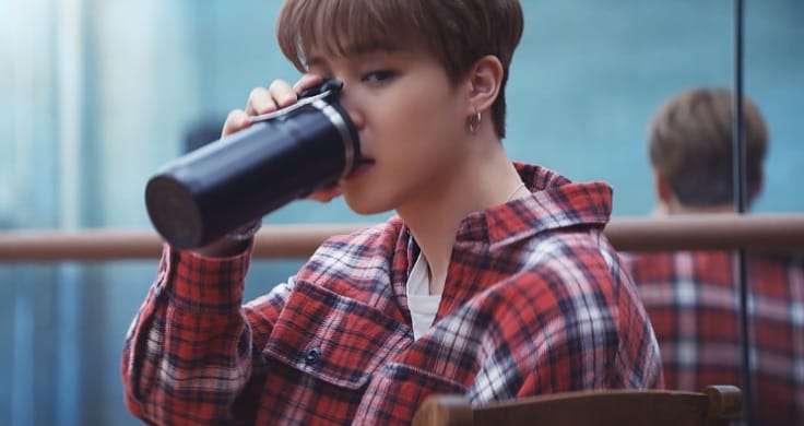 BTS member Jimin wearing a red, blue and white chequered flannel shirt takes a sip from a black reusable bottle.