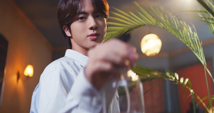 BTS member Jin wearing a white shirt and holding a clear bottle in with palm fronds behind him.