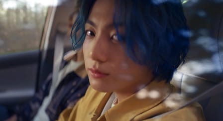 BTS member Jungkook sitting in a car with his seatbelt on next to an unseen person in a denim jacket.