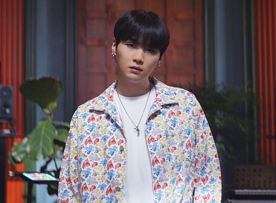 BTS member Suga wearing a colorful jacket stands in a room with house plants behind him. 