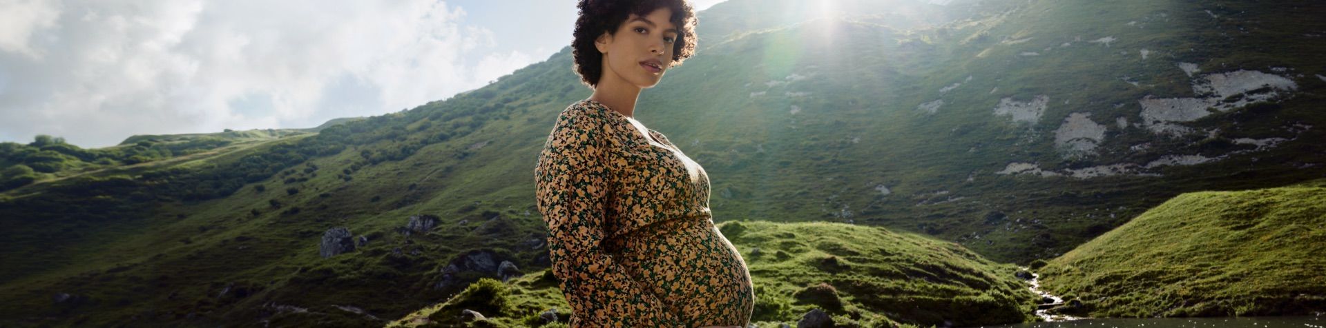 A pregnant woman in a yellow dress with a flowery print stands in front of mountains with the sun peeking through the clouds. 