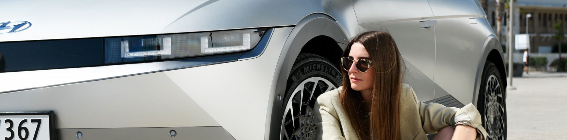 Britta Reineke sitting down in front of a gray IONIQ 5. She has long brunette hair and is wearing large tortoise shell sunglasses and a beige blazer. 