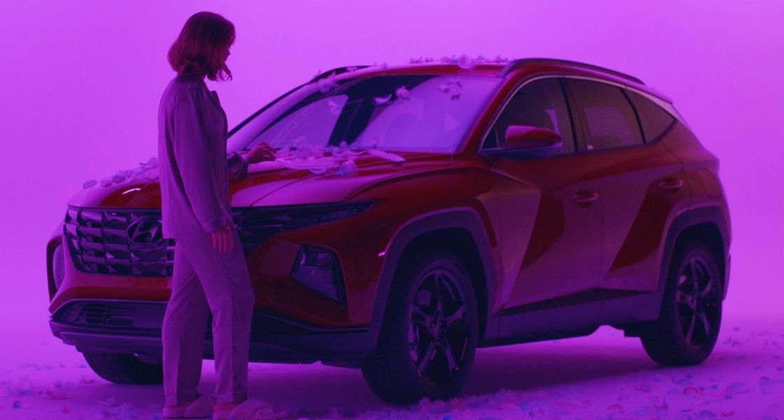 Side view of a red TUCSON plug-in hybrid covered in feathers and bathed in a purple light. ASMRtist Charlette Hastings is standing next to it and running a large white feather across the hood.
