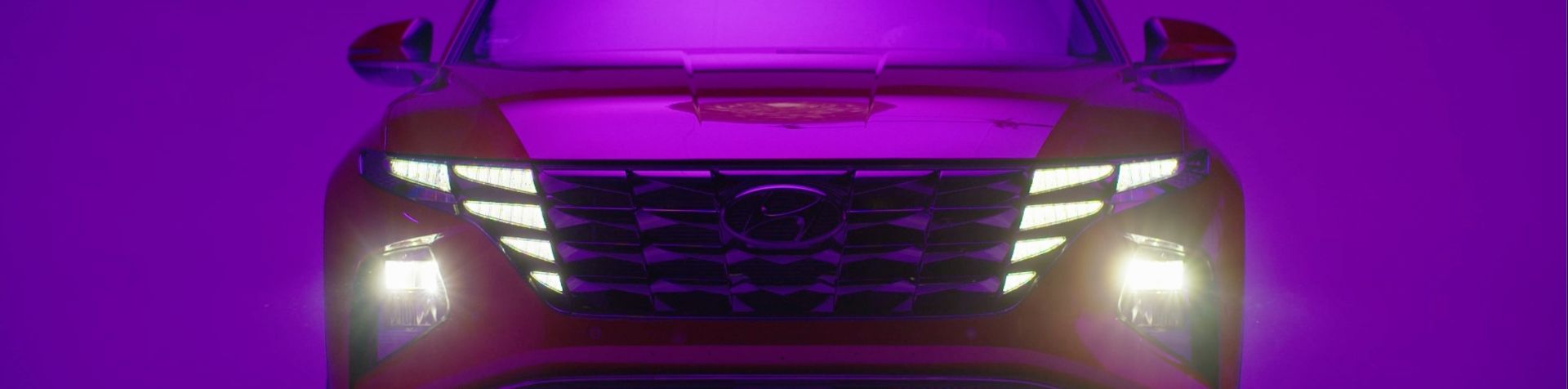 The front of a red TUCSON plug-in hybrid with its parametric headlights on against a purple background.