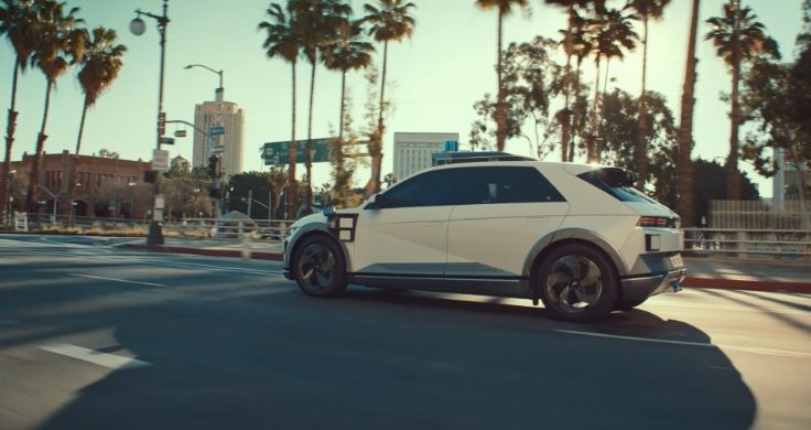 A white Hyundai IONIQ 5-based robotaxi driving along a road lined with palm trees in LA.