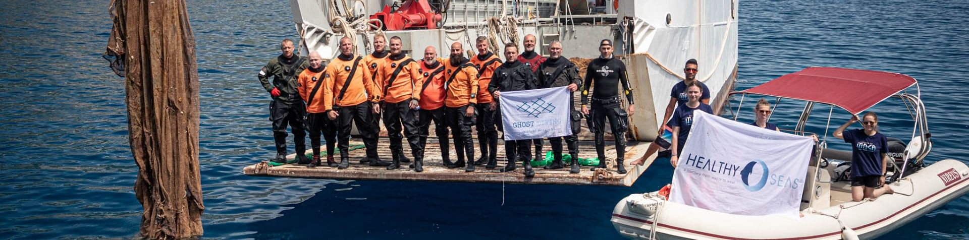 The ocean clean-up team dressed in orange standing on the back of a boat with a smaller boat next to it waving the Healthy Seas flag. 
