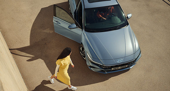 A woman wearing a yellow dress is passing in front of a car with an open door.