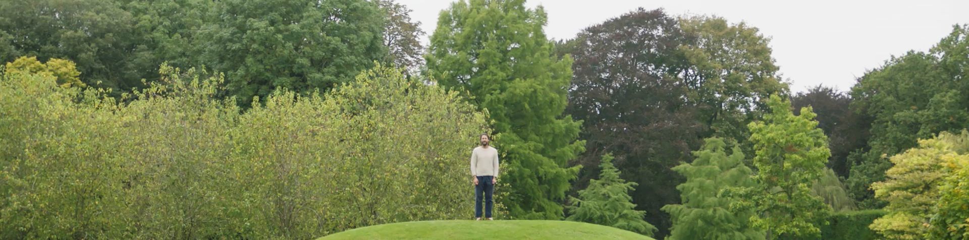 나무와 녹색 언덕에 서 있는 데이비드 드 로스차일드. 그는 맨발이고 회색 점퍼와 짙은 청바지를 입고 있습니다.
