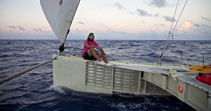 David de Rothschild sitting on his Plastiki boat in the middle of the ocean. He is wearing a pink t-shirt, black shorts and white sneakers and his hair is loose.   