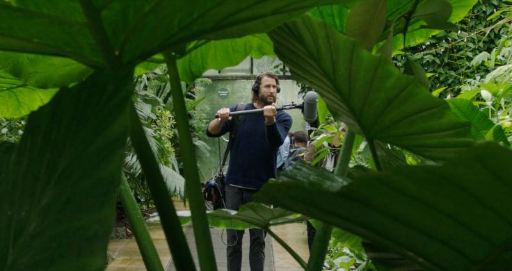 David de Rothschild holding a microphone up to some large green plants inside a greenhouse.