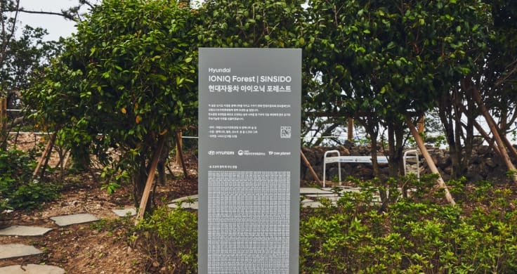 A gray sign at the opening of the IONIQ Forest Sinsido. Behind the sign a path winds through some trees to a white bench on the right-hand side.