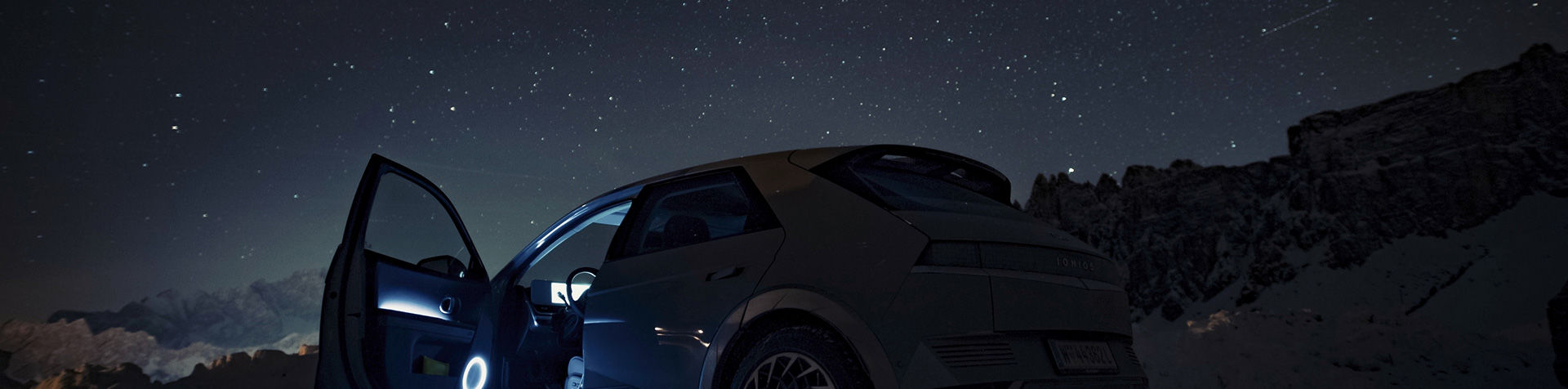 An IONIQ 5 standing with the side door open in the middle of a mountainous landscape with stars shining overhead.