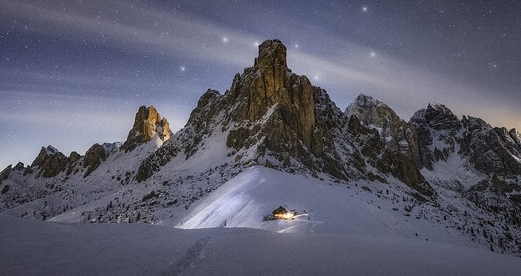 A snowy mountain with rocky outcrops with stars shining behind it.
