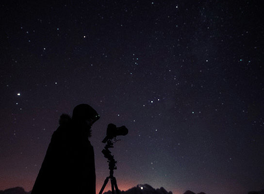 A silhouette of Nicholas Roemmelt looking through his camera at the night’s sky which is full of stars. 