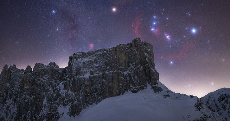 A mountain with a rocky outcrop covered in snow with a constellation of stars lit up behind it in the night sky. 