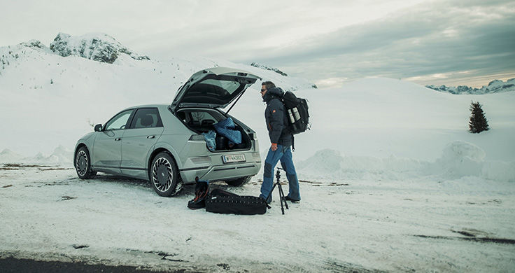Photographer Nicholas Roemmelt unloading his equipment from the trunk of a silver IONIQ 5 which is parked in the middle of a snowy and mountainous landscape.