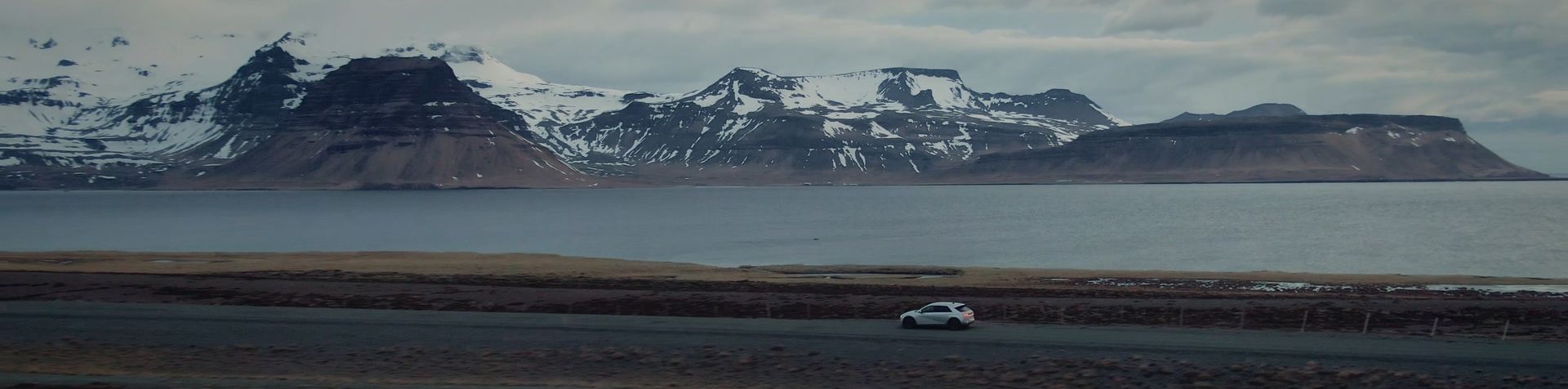 A gray IONIQ 5 drives along a road beside a lake with snow-capped mountains in the background.