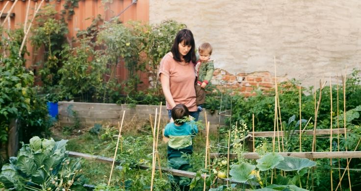 A mother holding one child in her arm and holding the hand of the other as they walk through a walled vegetable garden filled with green plants.