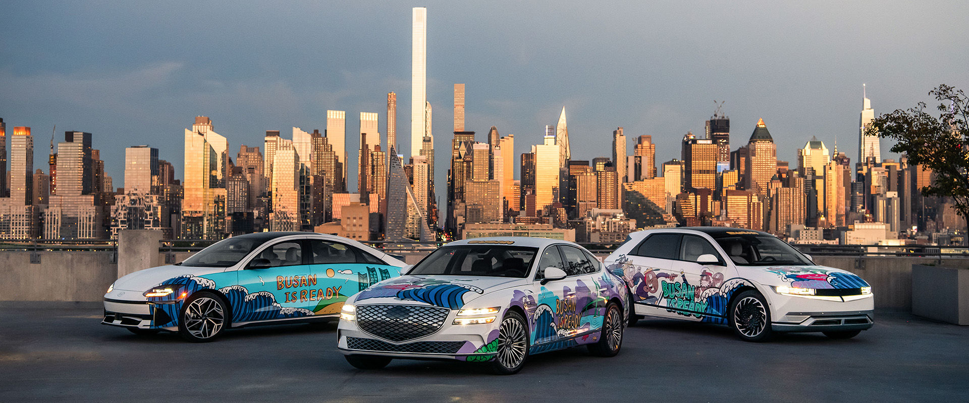 Genesis Electrified G80 sedan (center), Hyundai IONIQ 6 (left) and IONIQ 5 (right) show graffiti artwork and the slogan to promote Busan's bid to host the 2030 World Expo at Hudson County, New Jersey, with a New York City skyline in the background