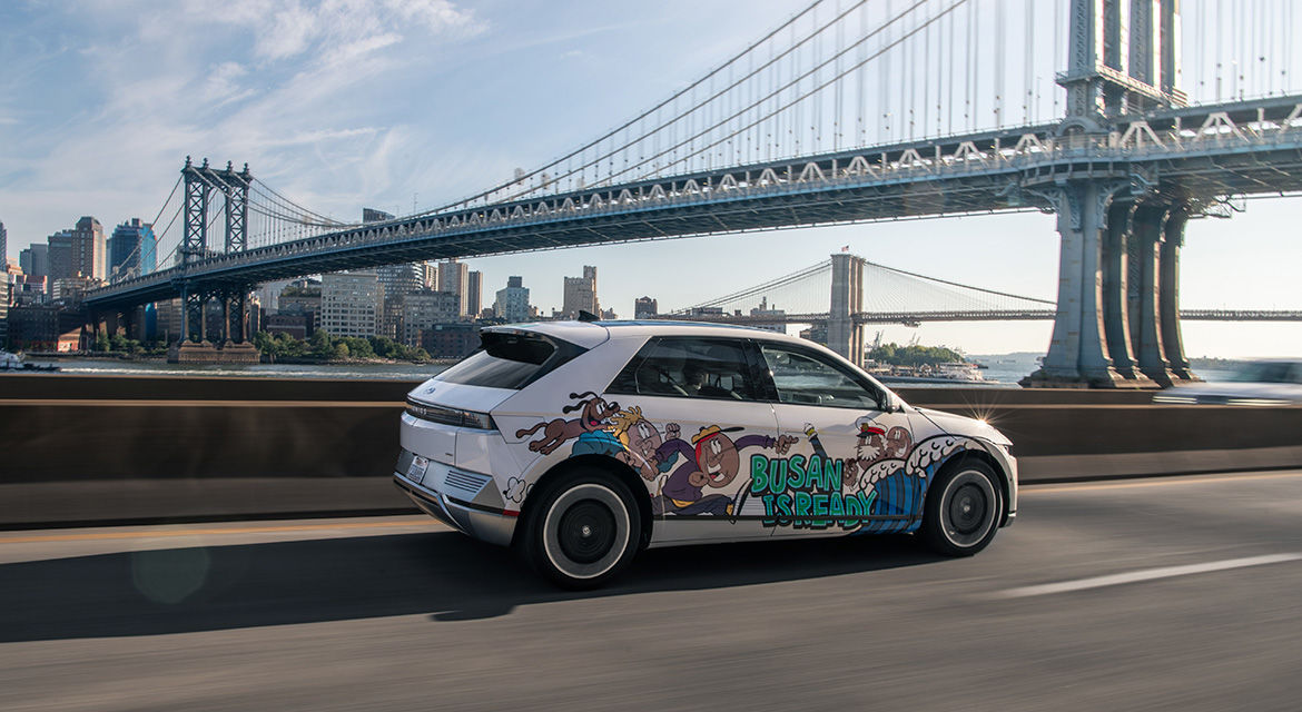 Hyundai's IONIQ 5 art car shows a side body wrapping that says "BUSAN Is Ready" driving along the Franklin D. Roosevelt East River Drive, New York, with the Manhattan Bridge and Brooklyn Bridge in the background