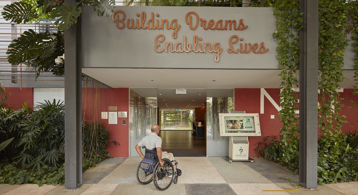 A man in a wheelchair is facing the entrance of the Enabling Village, the first inclusive community space in Singapore for persons with disabilities, managed by SG Enable. The signboard at the gate says "Building Dreams, Enabling Lives."