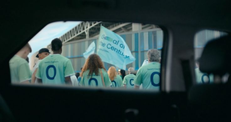 A view through a car window of a group of people of different ages all wearing Team Century jerseys and waving a flag with “Goal of the Century” on it. 