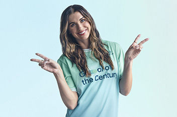 A close-up of Alex Morgan’s head and shoulders. She is wearing her green Team Century jersey and she has long wavy brunette hair. She is making the ‘v’ or peace sign with both hands.