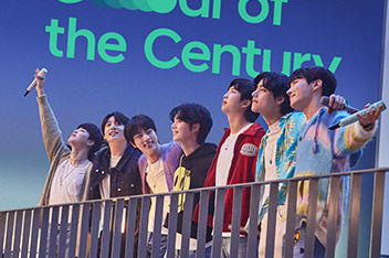 All 7 members of BTS on stage facing an unseen audience with 'Century' written in green on a blue screen behind them.