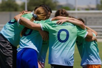 A group of children wearing green and blue Team Century jersey hug each other in a circle on the pitch.