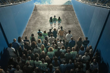 Bird’s eye view from behind a group of football fans walking through a corridor leading to the pitch. Three female footballers with long hair wearing green Team Century shirts with a blue number 0 on the back and white shorts and football socks and shoes are leading the group by a few steps.