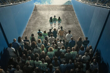 Bird’s eye view from behind a group of football fans walking through a corridor leading to the pitch. Three female footballers with long hair wearing green Team Century shirts with a blue number 0 on the back and white shorts and football socks and shoes are leading the group by a few steps.