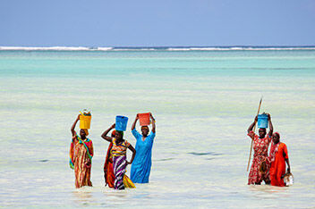 6 Senegalese women wearing brightly colored dresses and skirts carrying colorful buckets on their heads and wading through water.
